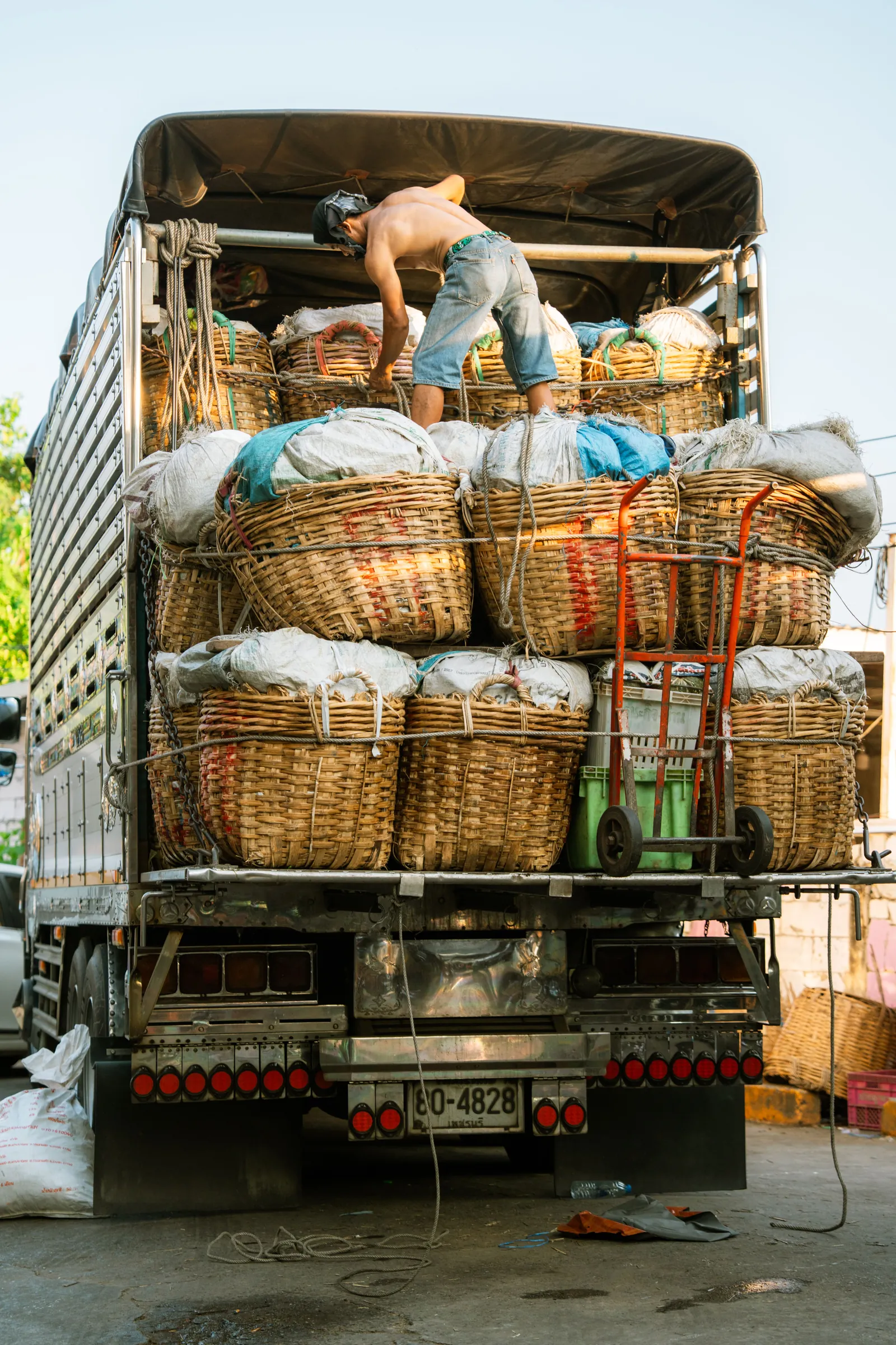 A worker standing on top of a truck loaded with woven produce baskets at a market in Bangkok