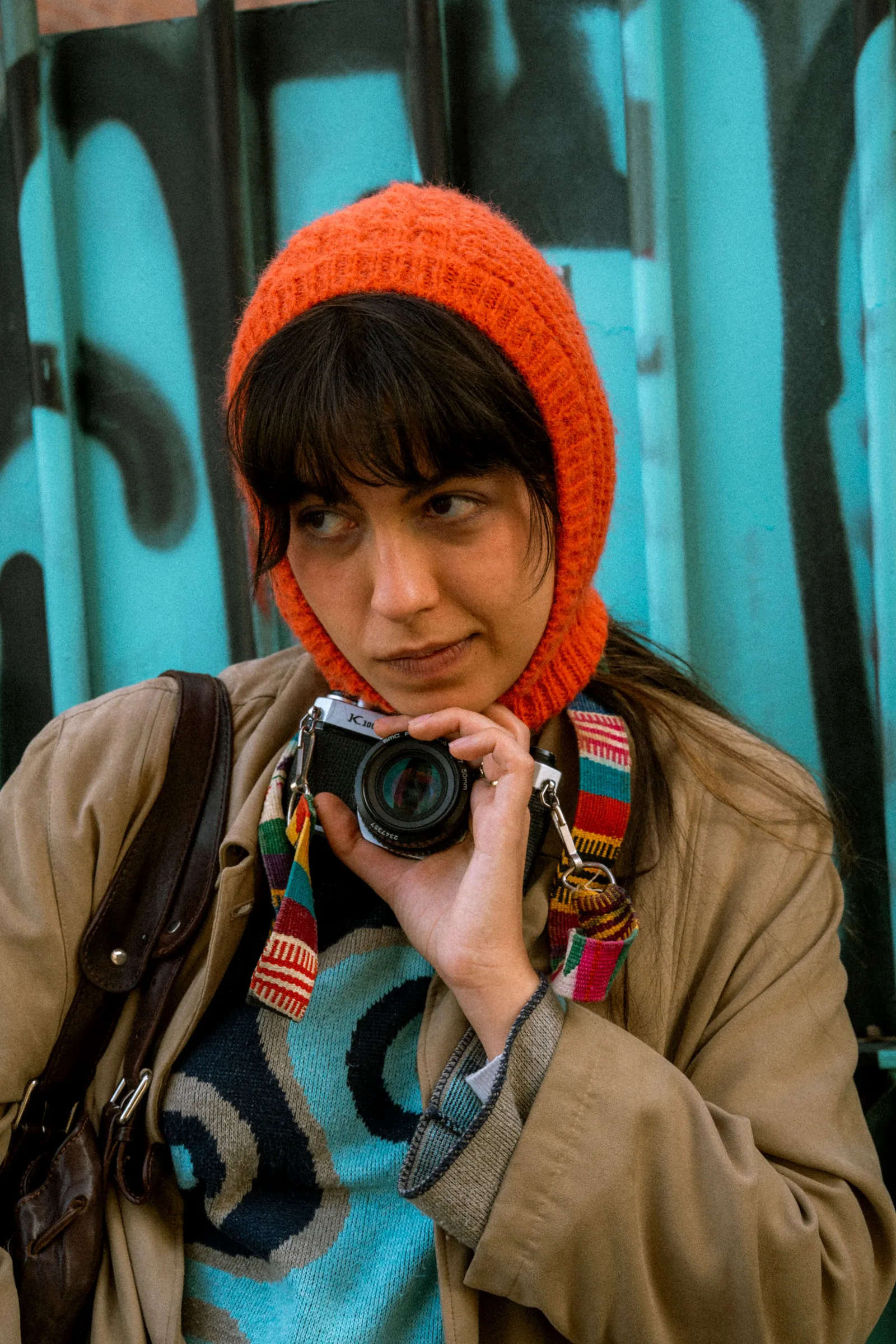 A young woman in an orange knitted hood holding a camera, standing in front of a turquoise fence covered in graffiti