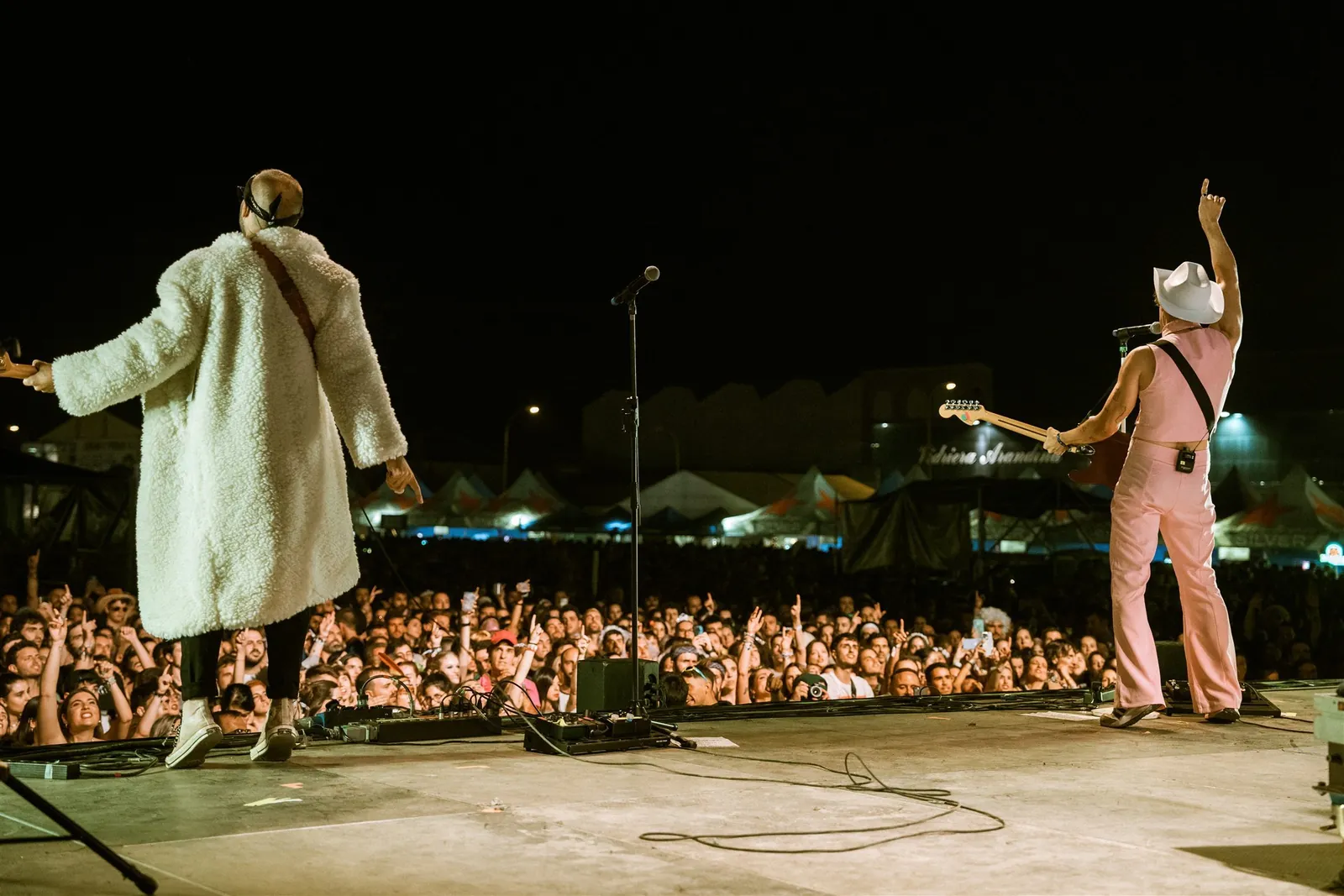 Two performers seen from behind on a festival stage, facing a large crowd at night
