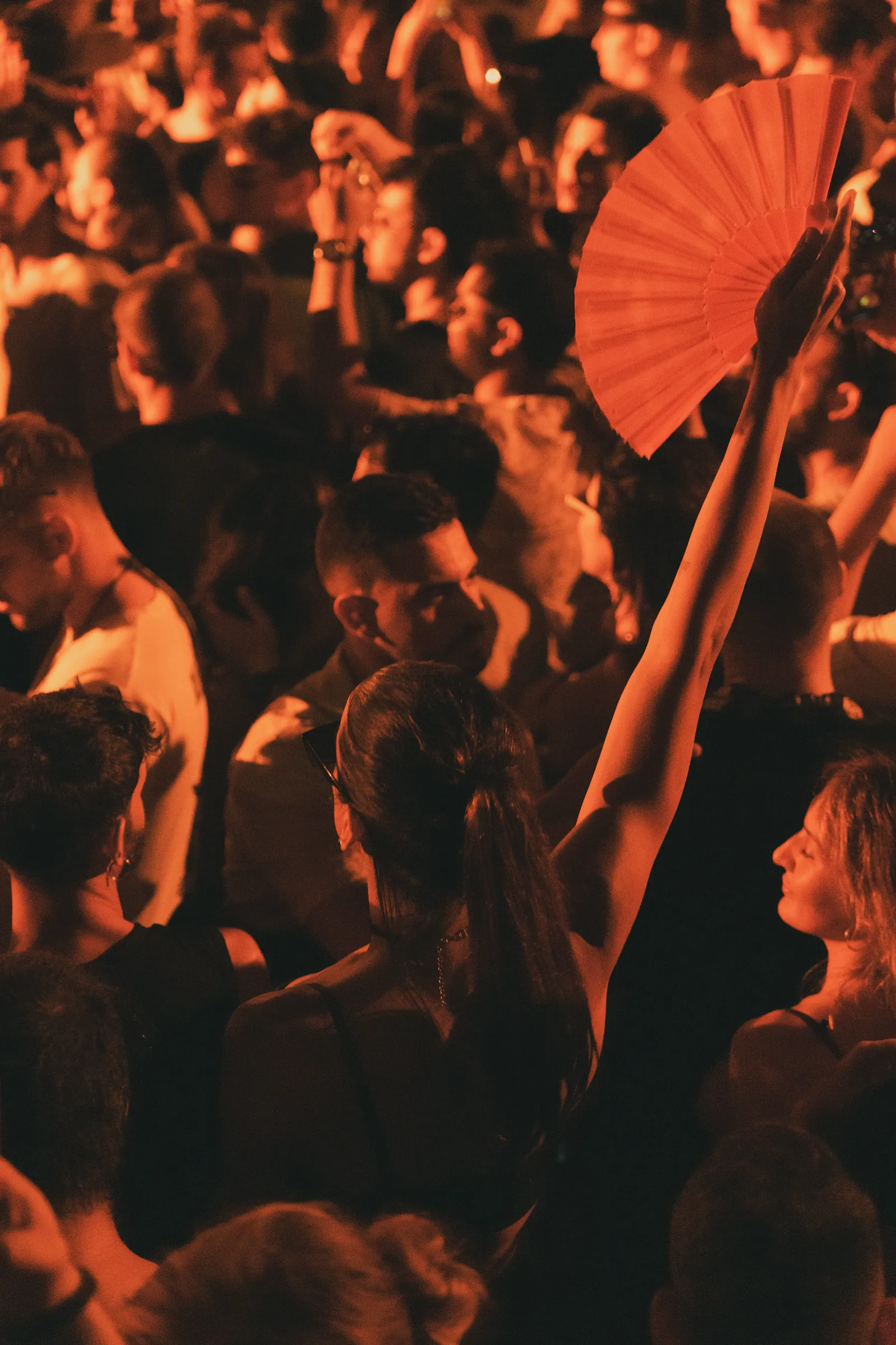 Festival crowd at night under warm orange light, with a raised arm holding a red hand fan above the heads