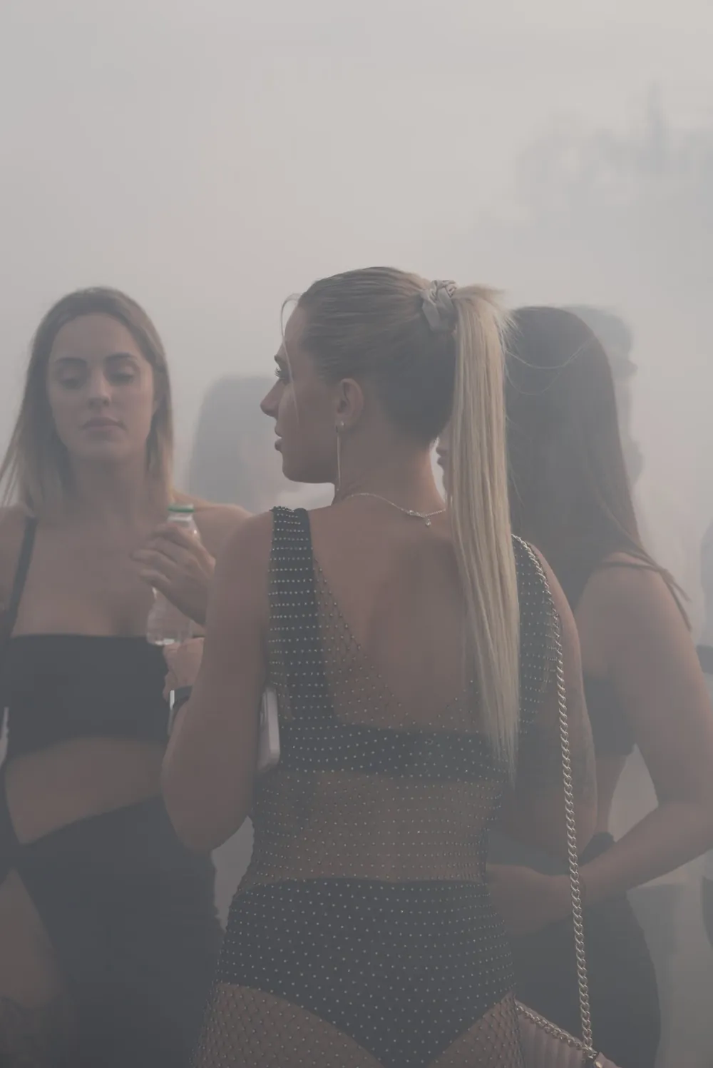 Unedited frame of three women talking inside thick festival smoke, with flat, bright, slightly pink colors