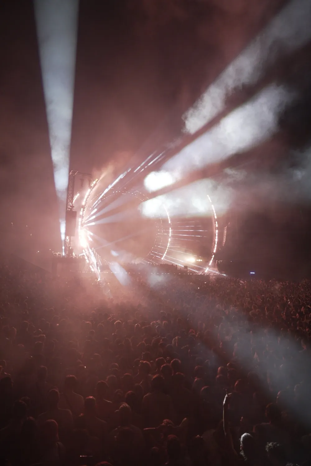 Unedited wide frame of a festival stage with light beams over a huge crowd, with a strong magenta-red cast