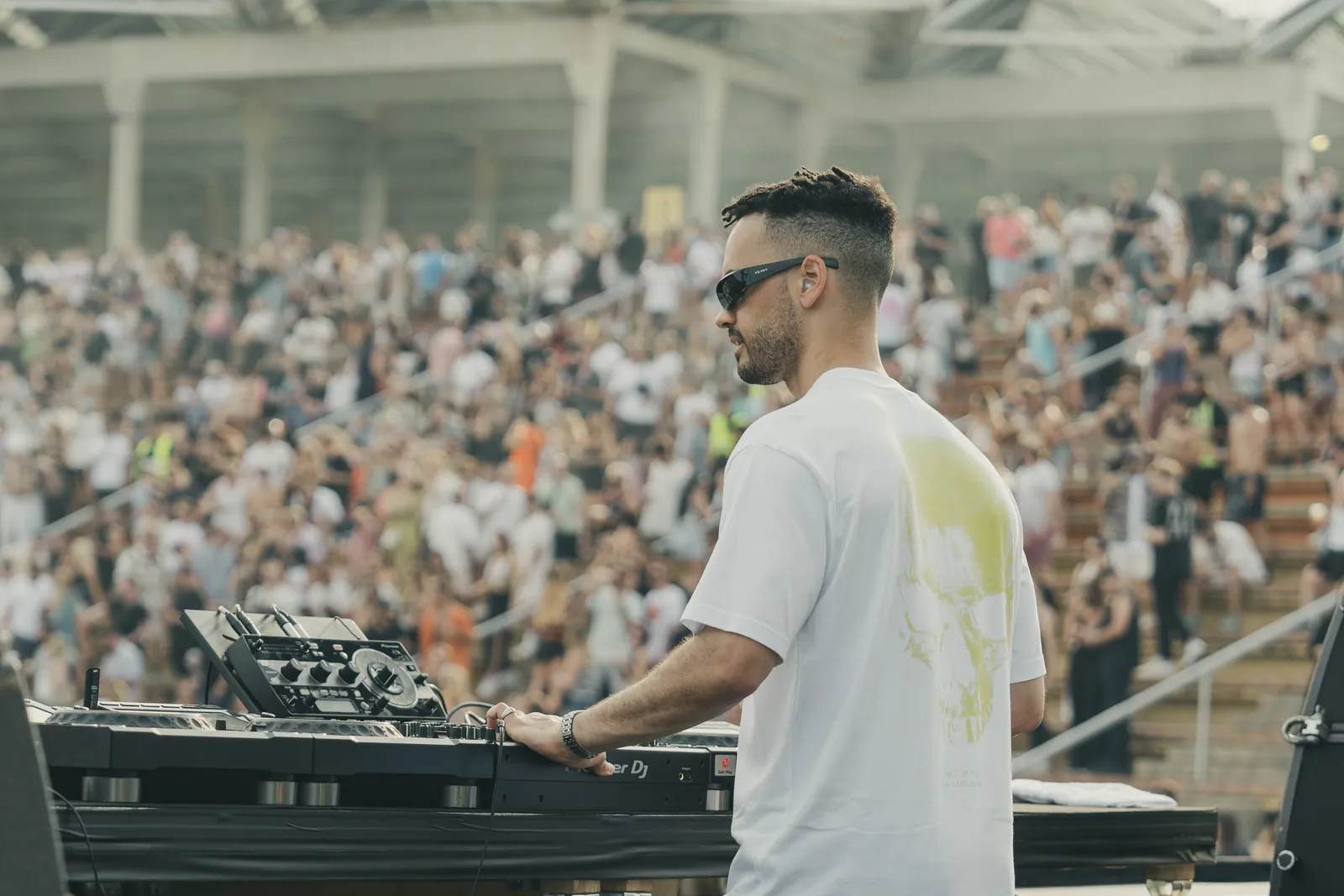 A DJ in a white T-shirt photographed from behind the decks, facing a packed festival crowd at OFF Week Festival in Barcelona